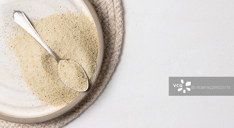 灰色盘子上的生未煮的丰年米种子，旁边放着一把金属勺子，白色桌面俯视图，非洲谷物。图片素材