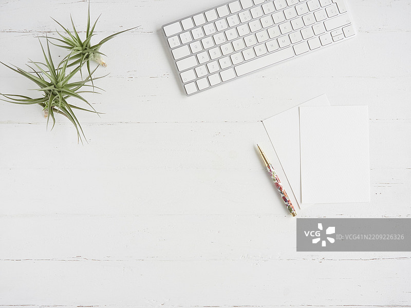 家用办公室的白色桌面平铺构图，留有空白空间。笔记本、空白纸张、钢笔、电脑键盘和绿色植物作为装饰。图片素材