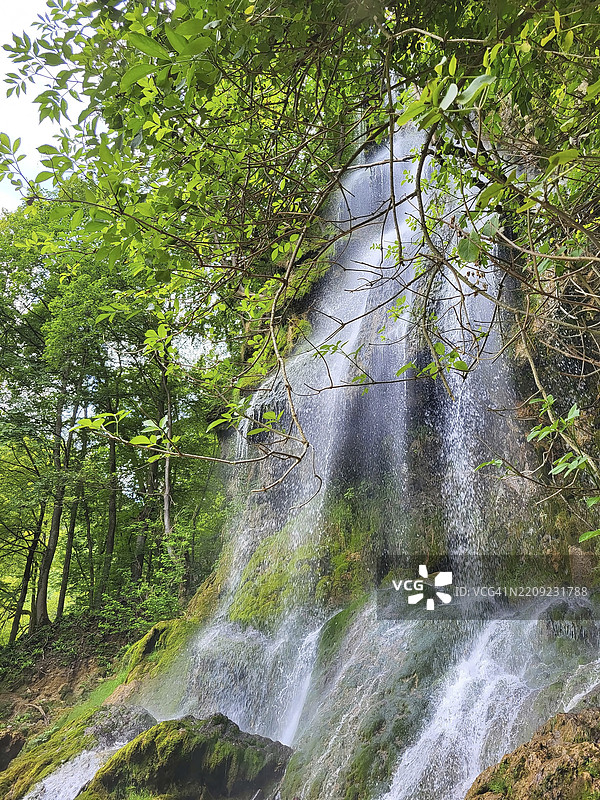 巴乌拉赫的瀑布，嵌入郁郁葱葱的森林景观中。水流从岩石面上倾泻而下，形成一个小水池，周围是覆盖着苔藓的石头和茂密的植被。阳光透过树冠洒下，营造出迷人的氛围。图片素材