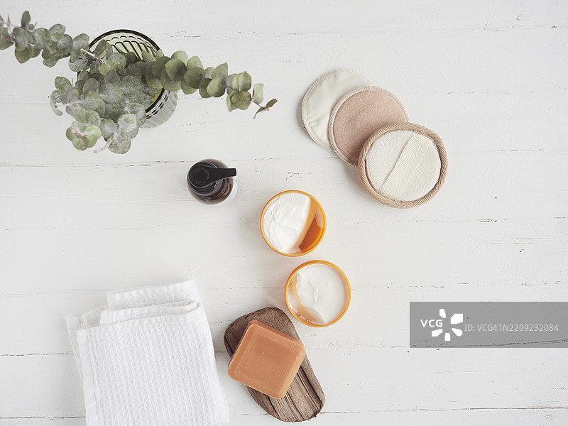 零废弃有机化妆品概念平铺组合，包括可重复使用的卸妆棉盘、有机肥皂、面霜、玻璃瓶、白色毛巾和干燥的桉树叶图片素材