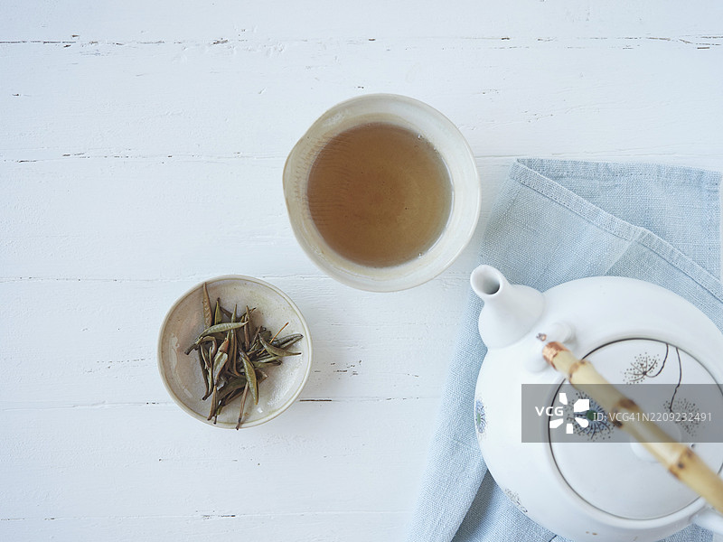 白色木桌上，蓝色布料旁边的茶壶旁，盛有银针白茶的茶杯，茶叶芽浸泡在碗中，热饮用瓷器盛装。俯视图。图片素材