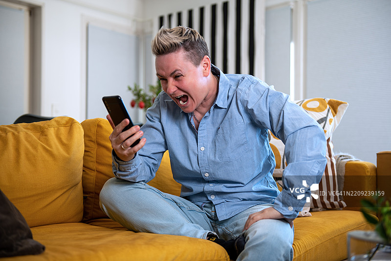 愤怒的人坐在家中黄色沙发上对着智能手机大喊。图片素材