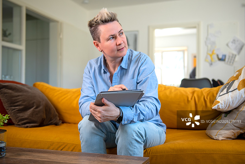 在舒适的家居环境中，坐在黄色沙发上的人使用数字平板电脑。图片素材