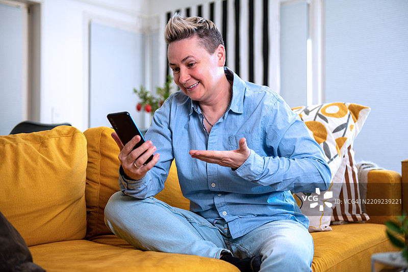在家中坐在黄色沙发上，愉快地通过智能手机视频聊天的人。图片素材