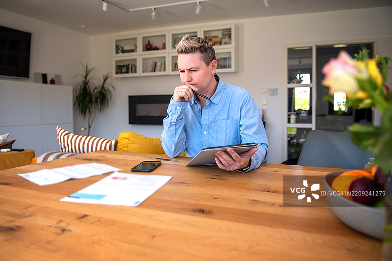 男子在日光明亮的家办公桌上审阅文件并使用平板电脑。图片素材