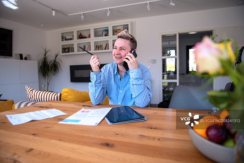 微笑的人在家中办公室桌子上打电话，桌上有文件。图片素材