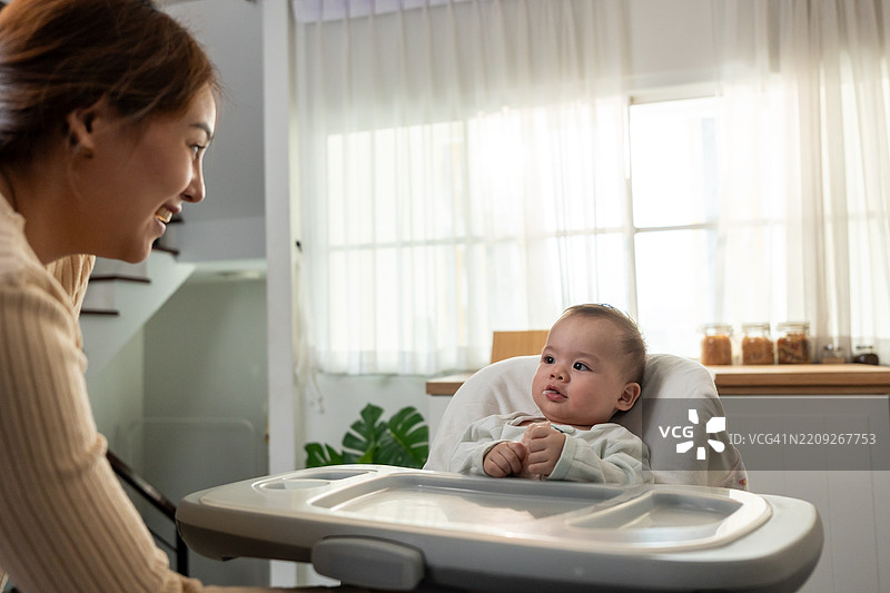 亚洲母亲在家中温柔地照顾和抚养婴儿男孩。迷人的妈妈在厨房里与坐在儿童椅上的新生儿分享温馨的时刻，营造出温暖的家庭氛围。图片素材