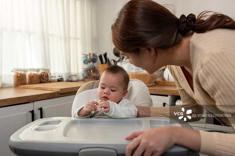 亚洲母亲在家中温柔地照顾和抚养婴儿男孩。迷人的妈妈在厨房里与坐在儿童椅上的新生儿分享温馨的时刻，营造出温暖的家庭氛围。图片素材