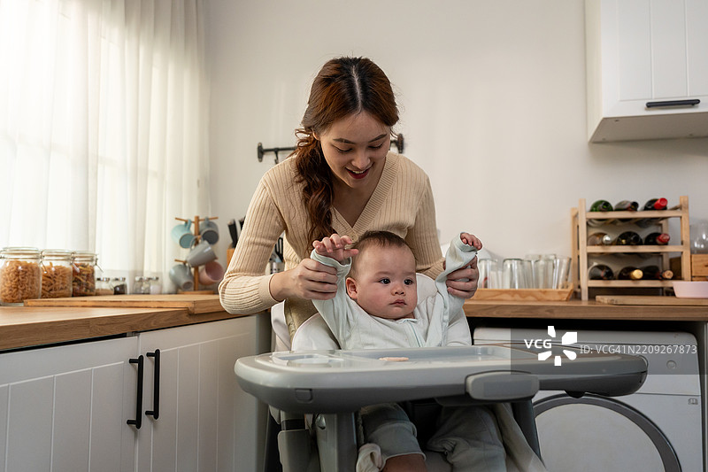 亚洲母亲在家中温柔地照顾和抚养婴儿男孩。迷人的妈妈在厨房里与坐在儿童椅上的新生儿分享温馨的时刻，营造出温暖的家庭氛围。图片素材