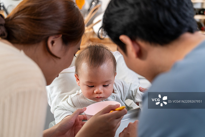 亚洲父母在家厨房里给小男孩喂健康食品。迷人的爸爸和妈妈温柔地照顾和抚养坐在儿童椅上的新生儿，营造出温馨的家庭氛围。图片素材