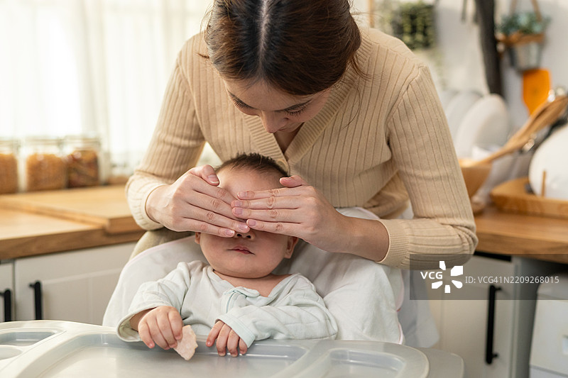 亚洲母亲在家中温柔地照顾和抚养婴儿男孩。迷人的妈妈在厨房里与坐在儿童椅上的新生儿分享温馨的时刻，营造出温暖的家庭氛围。图片素材