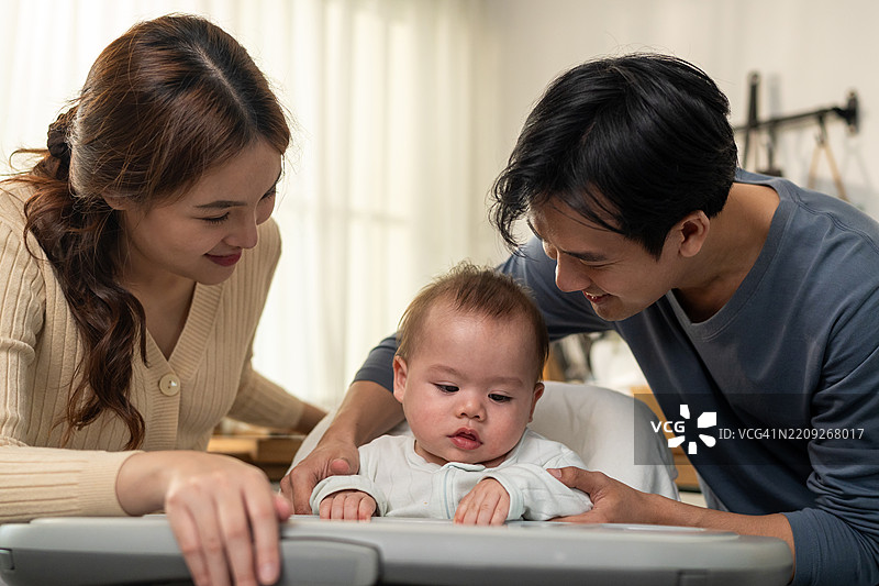 亚洲父母在家中温柔地照顾和抚养婴儿男孩。迷人的爸爸和妈妈在厨房里与坐在儿童椅上的新生儿分享温馨的时刻，营造出温暖的家庭氛围。图片素材