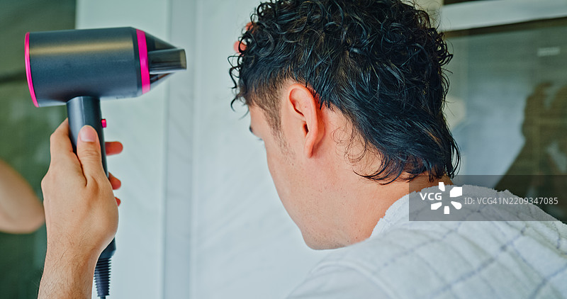 年轻男子在家浴室的镜子前吹干卷发。健康生活方式的早晨日常概念。图片素材