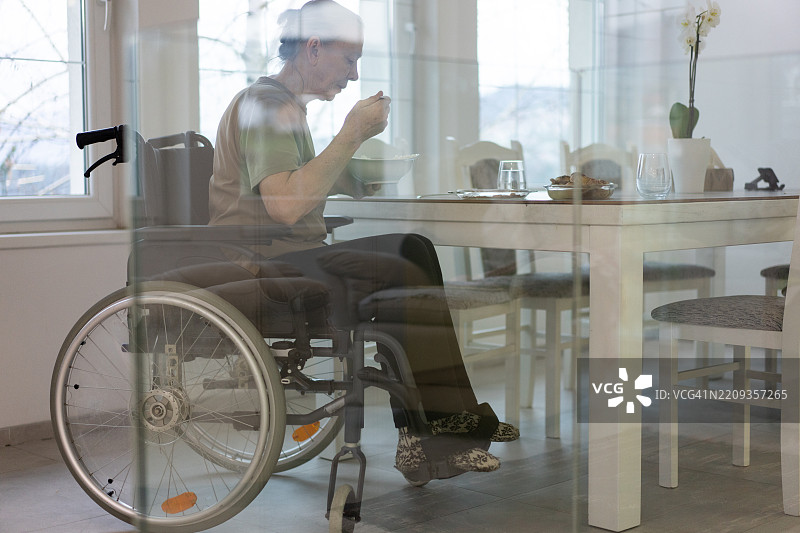 坐在明亮房间桌子旁的轮椅女性正在用餐。图片素材