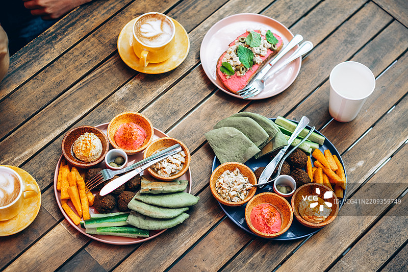 色彩鲜艳的素食菜肴平铺在古朴的木桌上。食物包括绿色皮塔饼、鹰嘴豆饼、鹰嘴豆泥、甜菜酱、黄瓜和红薯薯条。几只手伸入画面，营造出一种社交共享的用餐氛围。图片素材