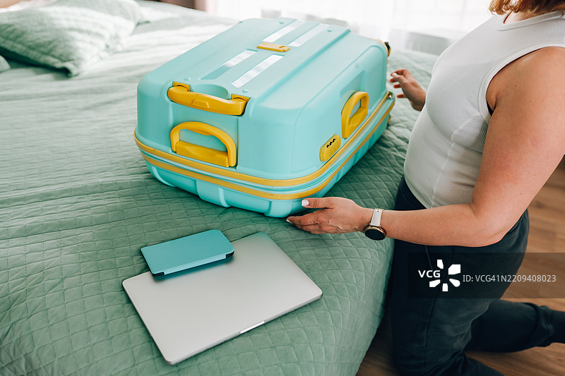 女人在床上打包浅蓝色行李箱，旁边有笔记本电脑和智能手机。图片素材