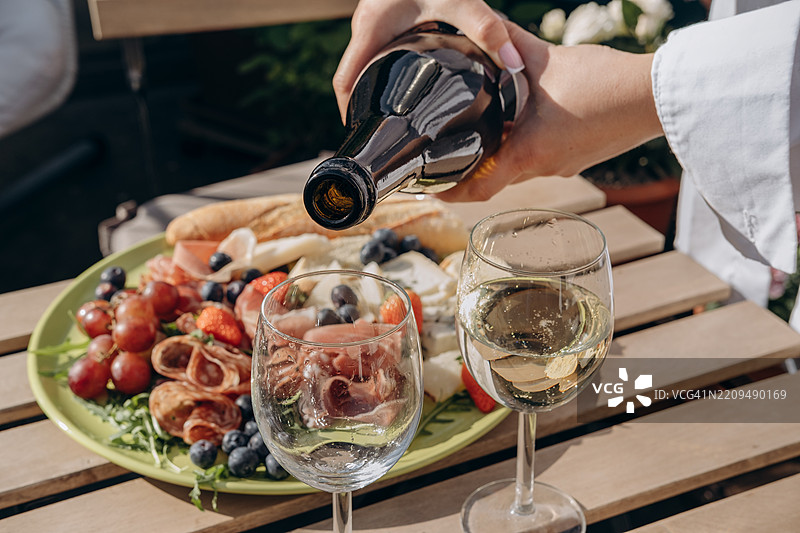 露台上的开胃酒，一位女士将白葡萄酒或普罗塞克酒从瓶中倒入酒杯，桌上摆放着一盘开胃小吃，包括生火腿、萨拉米香肠、戈尔根朱拉奶酪、帕尔马奶酪和浆果。图片素材