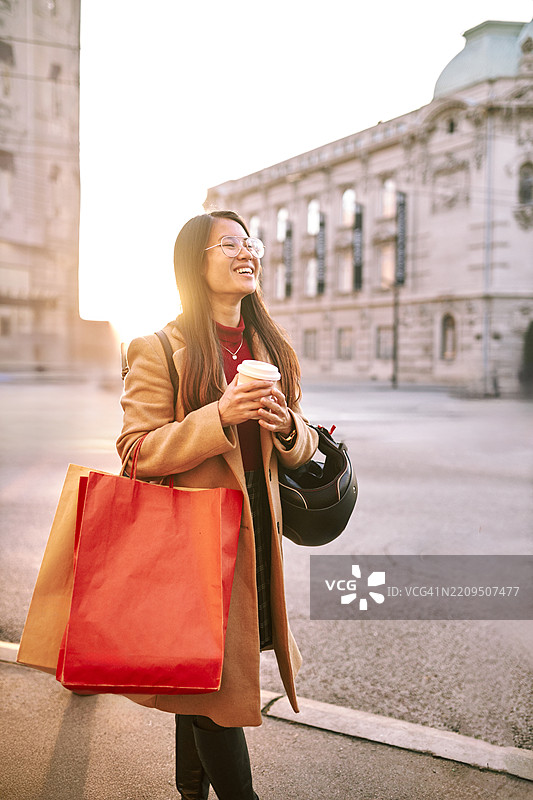 年轻时尚的女性在阳光明媚的城市街道上手握咖啡杯和购物袋。图片素材