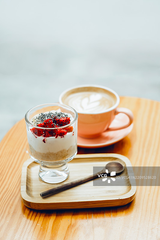 分层酸奶、水果和奇亚籽的玻璃杯，旁边是一个木托盘上的拿铁咖啡杯和勺子。图片素材