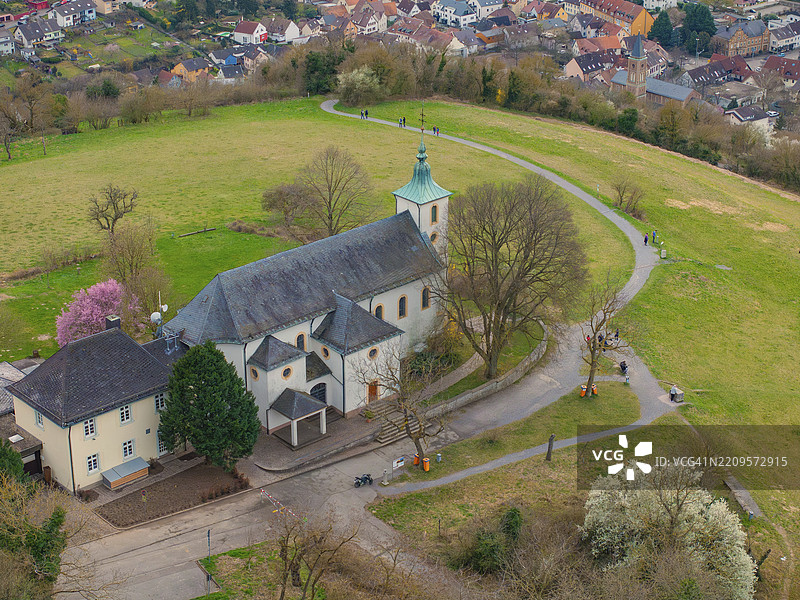 教堂建筑位于山坡小径上，四周环绕着春天的树木，背景是宁静的村庄，米歇尔斯贝格，巴德·门斯特赖费尔图片素材