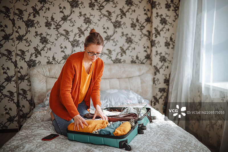 穿着温暖开衫的女性在舒适的床上将衣物折叠进行李箱，准备慢旅行。图片素材