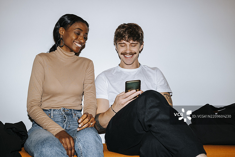 快乐的年轻男子坐在沙发上，使用智能手机，身边有一位男性朋友，靠近白色墙壁，正在上大学。图片素材