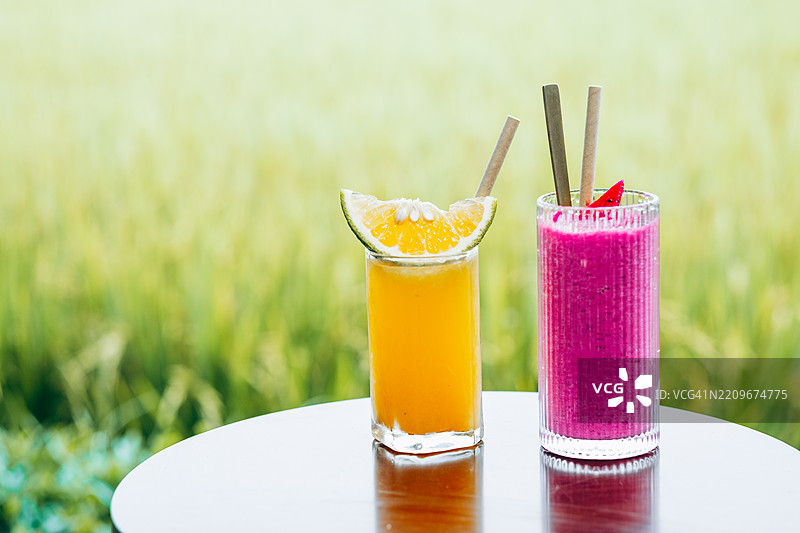 两杯色彩鲜艳的饮料，一杯橙色，一杯红色，放在桌子上，背景是模糊的稻田。图片素材