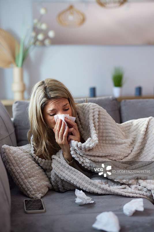 生病的女人在家里用毯子裹着自己，正在擤鼻涕。图片素材
