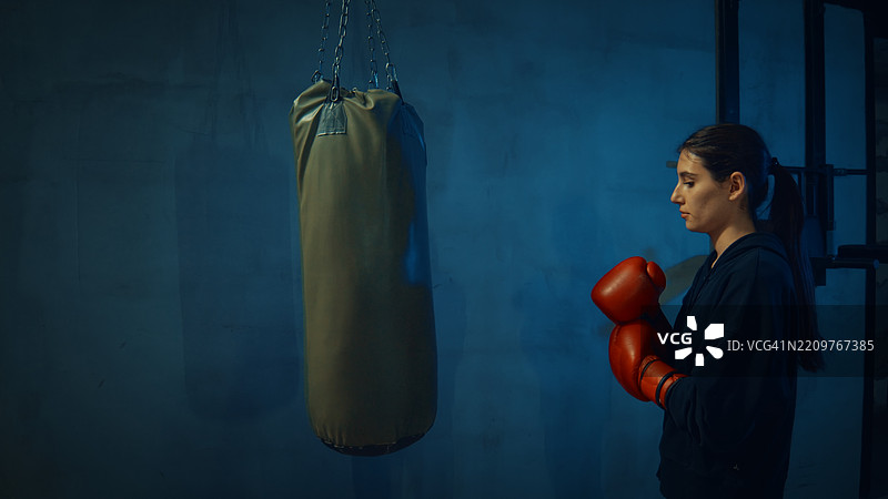 年轻女性拳击手戴着红色拳击手套，注视着黑暗健身房中的沙袋图片素材