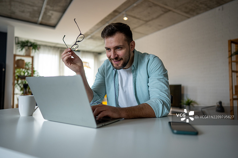 年轻男子在家用笔记本电脑工作，手里拿着眼镜图片素材