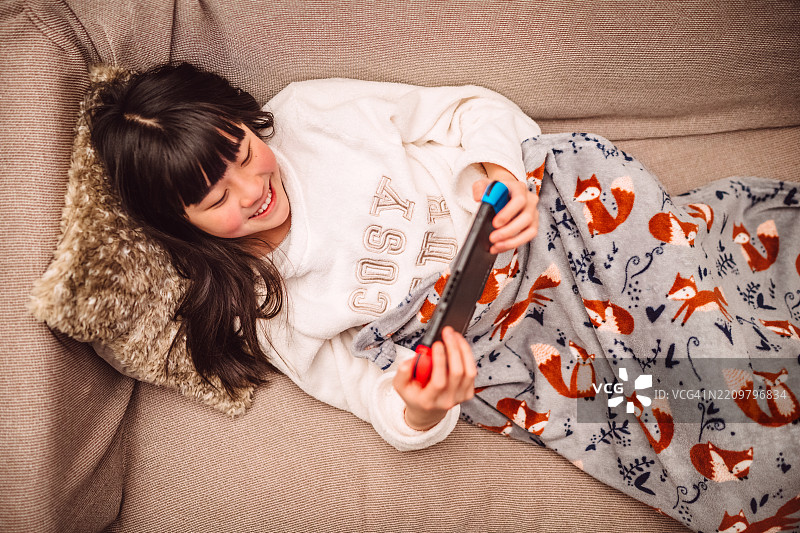 从上方直接俯视一名青少年女孩在家中放松，坐在沙发上玩视频游戏。图片素材