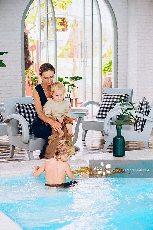 女人和金发幼儿坐在泳池边。另一个金发孩子在水中玩耍。周围环境明亮，绿植环绕，光线柔和。图片素材