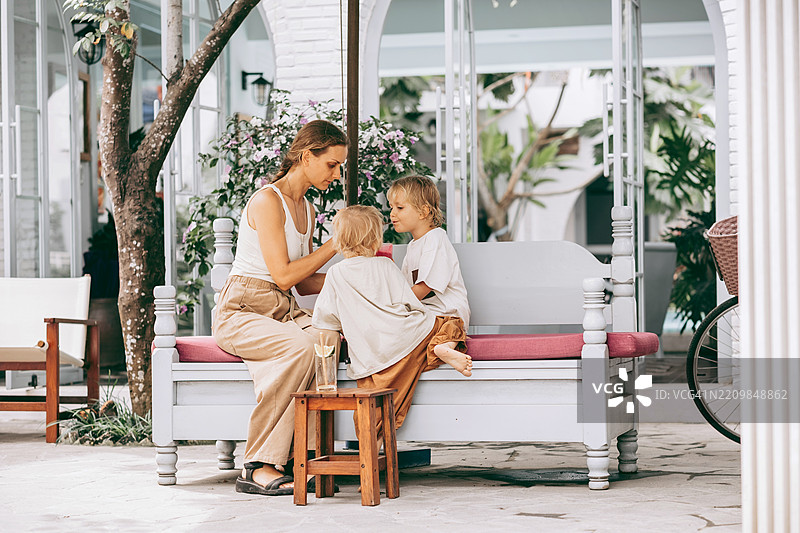 母亲坐在户外的白色长椅上，身边有两个金发孩子。她一边喂其中一个孩子，一边在粉色靠垫和小木凳旁放松。图片素材