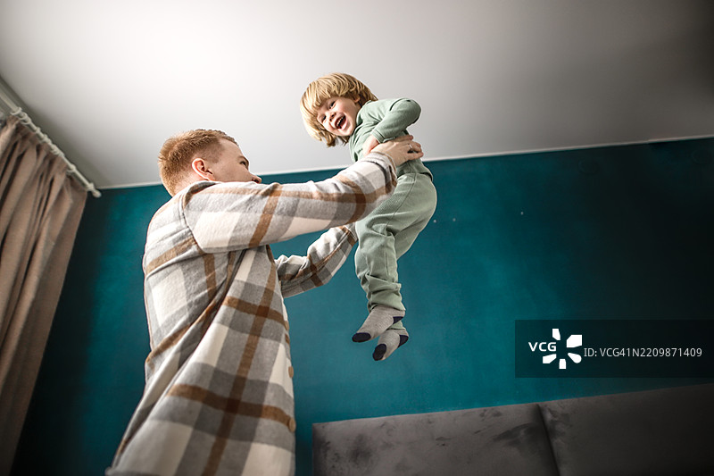 父亲在温馨昏暗的卧室里支持儿子在床上跳跃，享受玩耍的时刻。图片素材