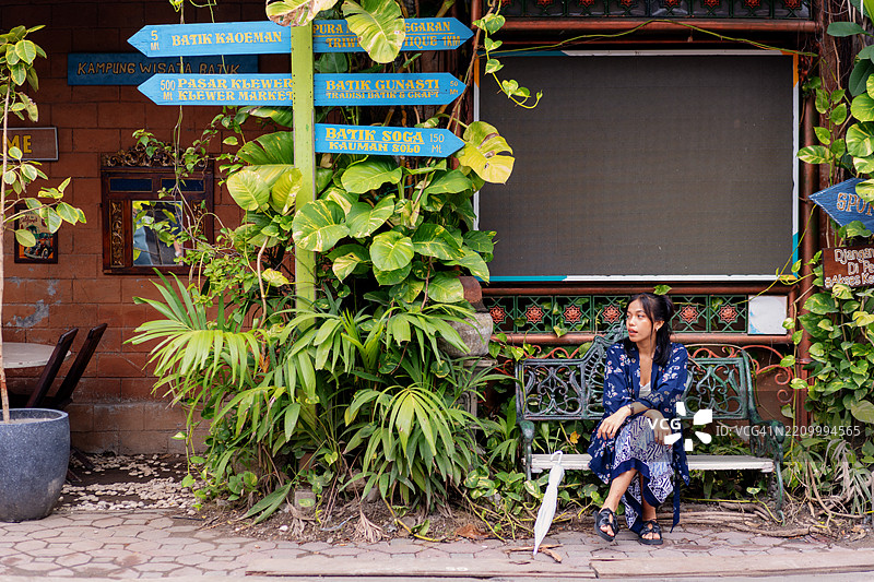 印尼女性坐在旅游村的公共长椅上，周围是指示牌和热带植物。图片素材