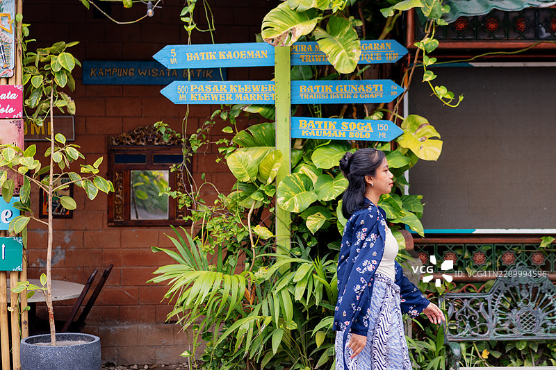 印尼女性在传统巴蒂克旅游村中漫步，周围是指示牌和热带植物。图片素材