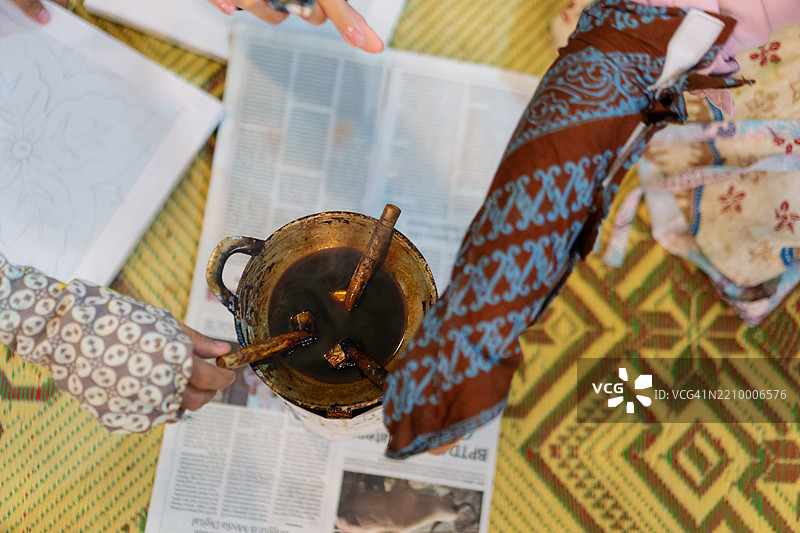 从上方俯视穿着保护性巴蒂克袖子的手，将浸蜡工具浸入融化的蜡中图片素材