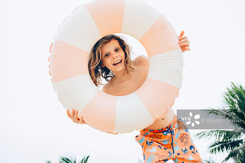 年轻男孩手持粉色充气浮圈，调皮地微笑，面前遮住了脸。穿着鲜艳的橙色泳裤，上面有海洋生物图案。背景是棕榈树。图片素材