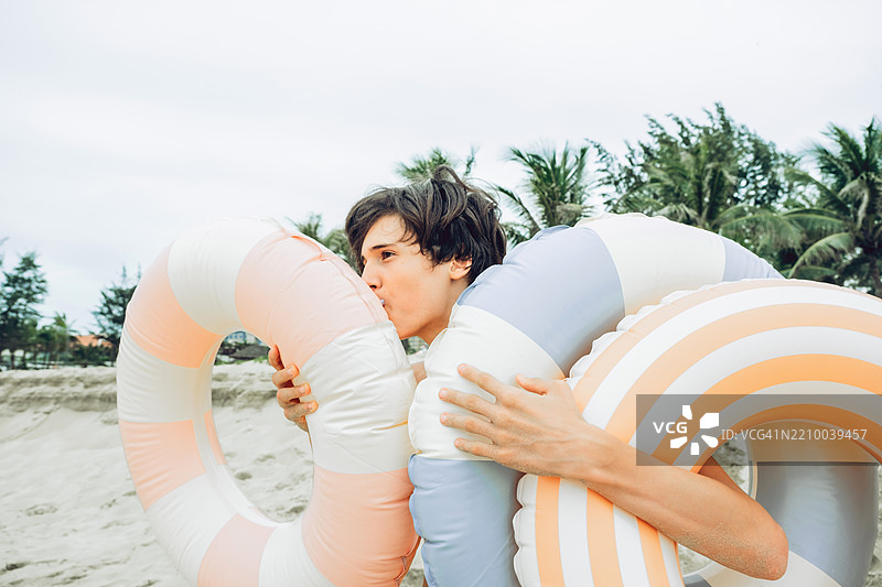 短发男孩在沙滩上充气五彩条纹的游泳圈。背景是棕榈树和多云的天空。穿着休闲的海滩服装，心情愉快。图片素材