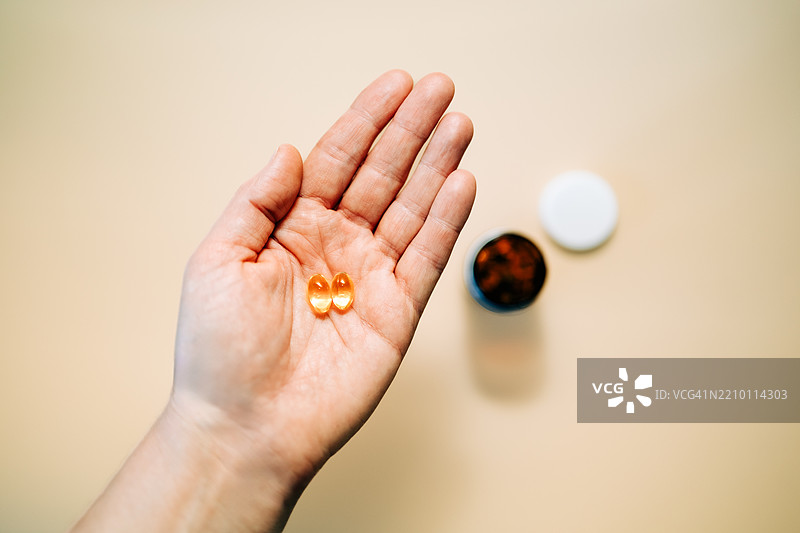 手握两个油胶囊，背景中有打开的药瓶图片素材