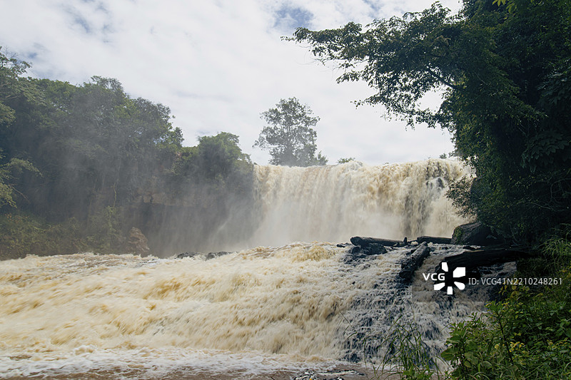 柬埔寨蒙多基里省的布斯拉瀑布，瀑布水流倾泻而下，雾气弥漫，周围绿植茂盛，天空阴云密布。图片素材