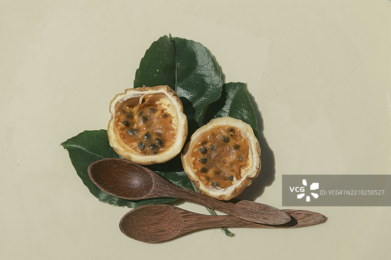 两半百香果，里面有种子，放在绿色叶子上，旁边有木勺，背景为米色。图片素材
