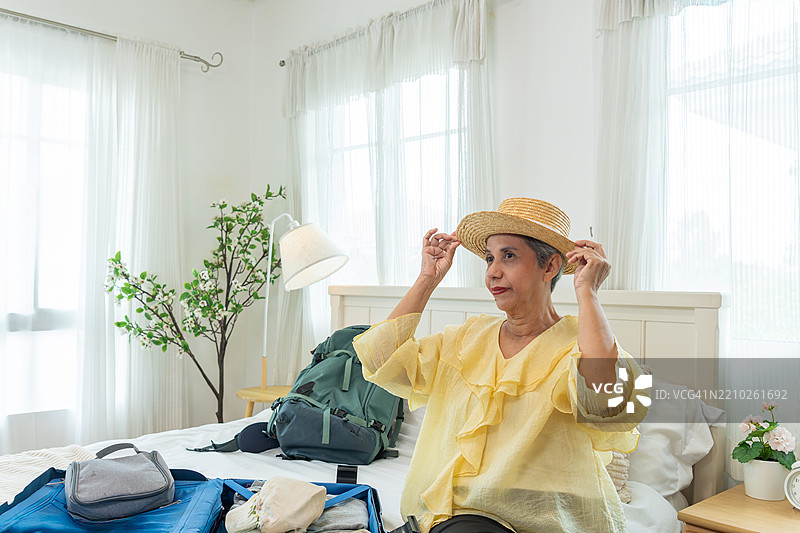 一位年长的亚洲女性在准备独自旅行，试戴帽子时正在打包。图片素材