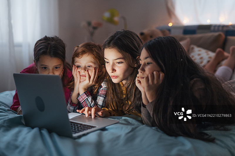 女孩们在床上躺着，通过笔记本电脑观看电影，享受朋友的睡衣派对。图片素材