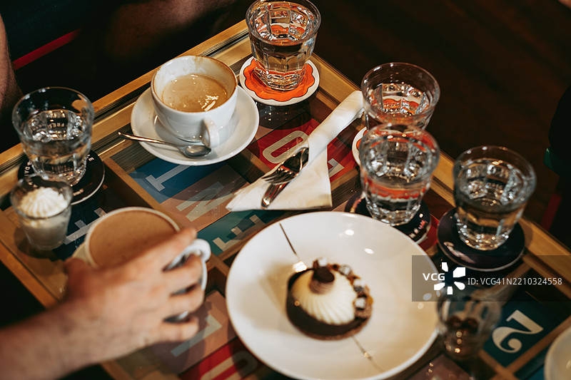 咖啡杯、小甜点和水杯摆放在色彩斑斓的咖啡桌上。一个人的手正伸向一个杯子。这个场景展现了一个轻松的室内咖啡馆环境。自然光线，温暖的色调。图片素材