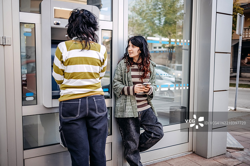 少女在户外的ATM机前取款，使用信用卡，而她的朋友在一旁等候。图片素材
