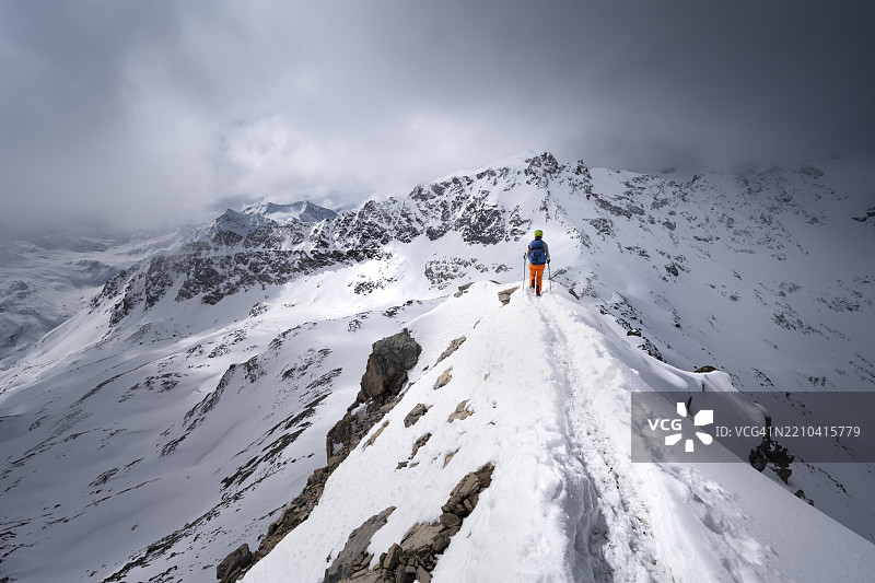 登山者在皮茨·拉维纳尔的狭窄山脊上下降,冬季山景中壮丽的山脉全景,伴随戏剧性的云彩,格劳宾登高路线,阿尔布拉阿尔卑斯山,瑞士莱蒂安阿尔卑斯山,格劳宾登,瑞士东部,欧洲图片素材