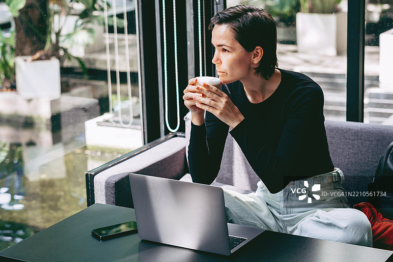 年轻的亚洲女性短发，坐在现代咖啡馆里，手里拿着咖啡杯，望着窗外。桌上放着一台笔记本电脑和一部智能手机。自然光线和窗外的绿植。图片素材