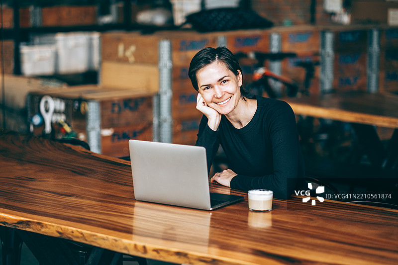 年轻的亚洲女性短发微笑着看向镜头，坐在长木桌前，桌上有笔记本电脑和咖啡。背景中有箱子和工具。图片素材