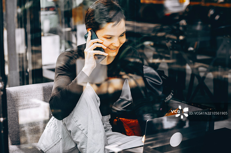 年轻的亚洲女性坐在咖啡馆的包厢里，微笑着用智能手机通话。玻璃墙上的倒影显示出植物、交通和城市细节。桌子上有一台笔记本电脑。概念：图片素材
